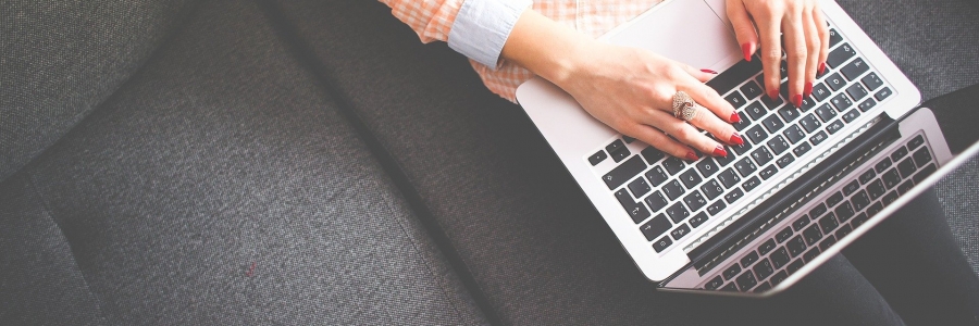 A person wearing an orange and white gingham patterned shirt works on a laptop while seated on a gray couch