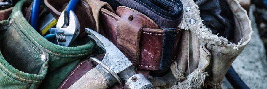 A worn tool belt is pictured laying on a grey pipe with a hammer laying on top and adjustable wrench emergig from the pocket of the belt.wrench  