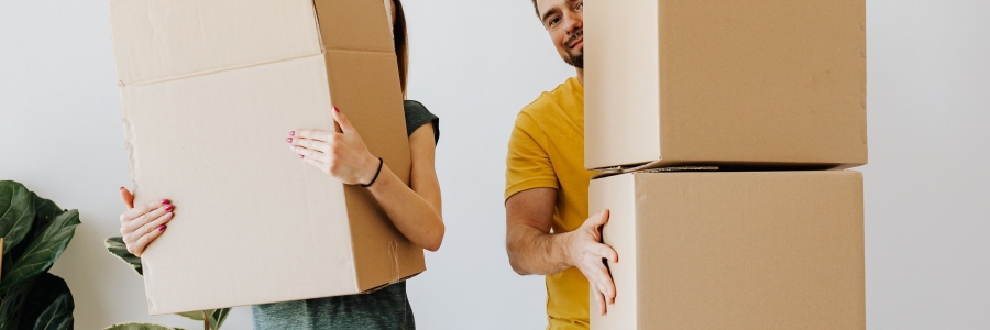 two peope in blue jeans are holding cardboard boxes and standing in front of a white wall with some plants on the left.