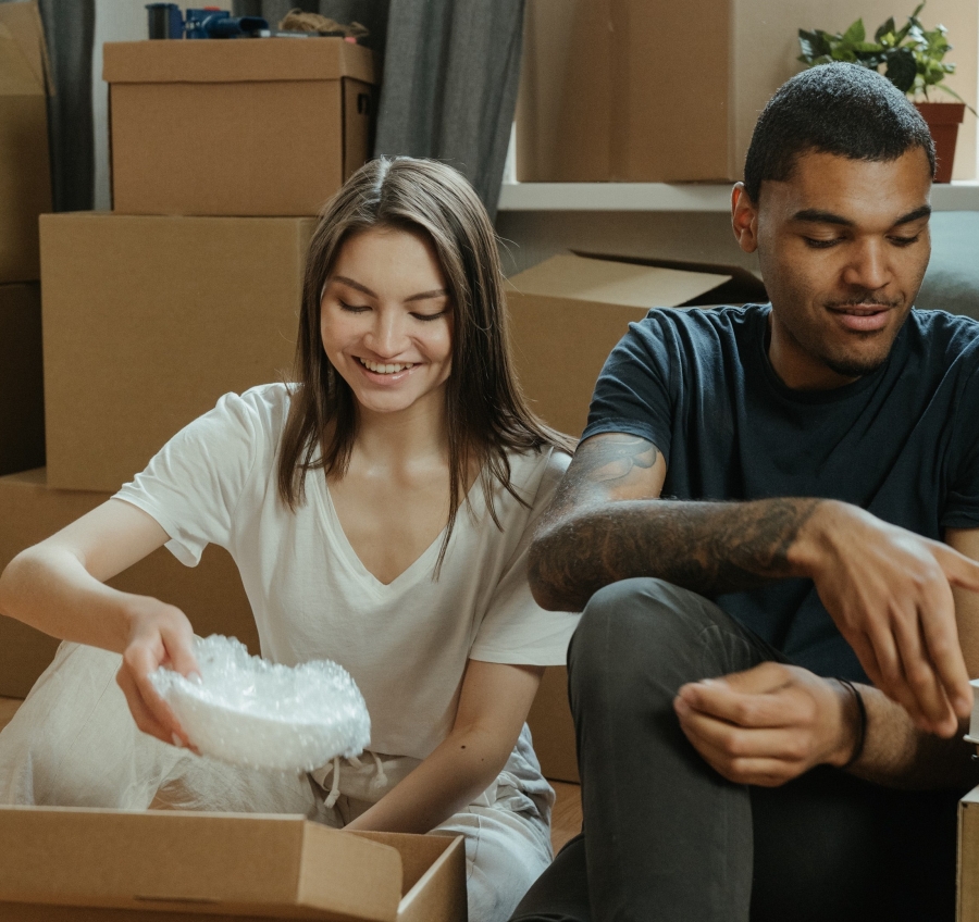 a man in a blue t-shirt and a woman in a white t-shirt sit among cardboard boxes. The woman is unpacking a white bowl wrapped in bubble wrap.