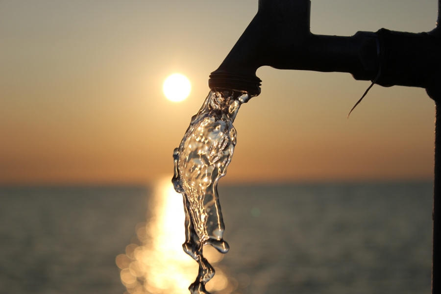 A running faucet is pictured with a sunsetting over a lake in the background.