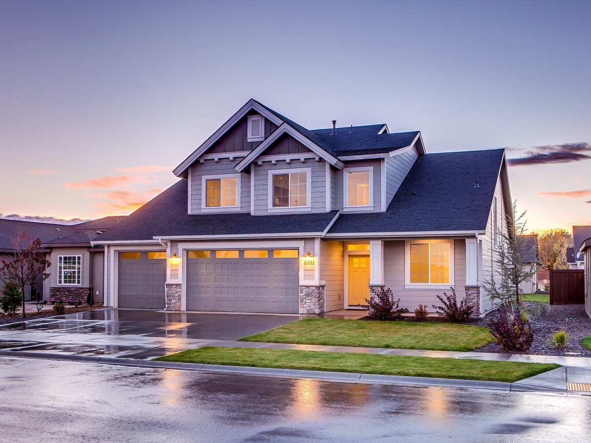 A gray house with black shingles is pictured with lights. The pavement looks wet as if it has just rained.