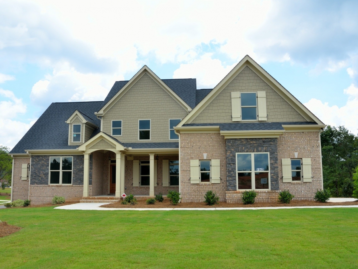 A beige brick home with a bright green lawn sits with a backdrop of trees and a blue sky with fluffy clouds.