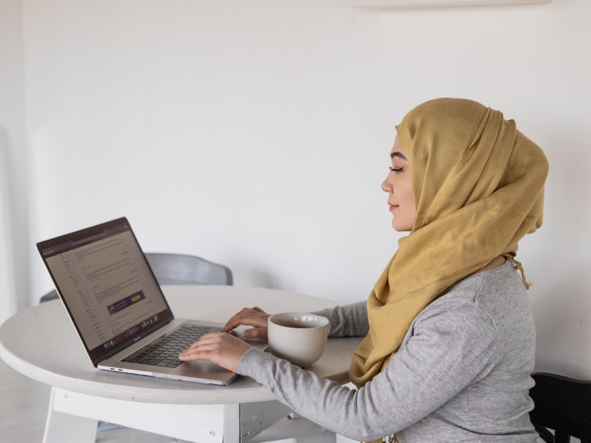 A woman wearing a mustard yellow hijab is seated at a table typing on a laptop.